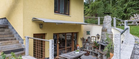 Edificio de dos pisos con fachada amarilla, una escalera de madera, un balcón con barandilla metálica y un muro de piedra con chimenea.