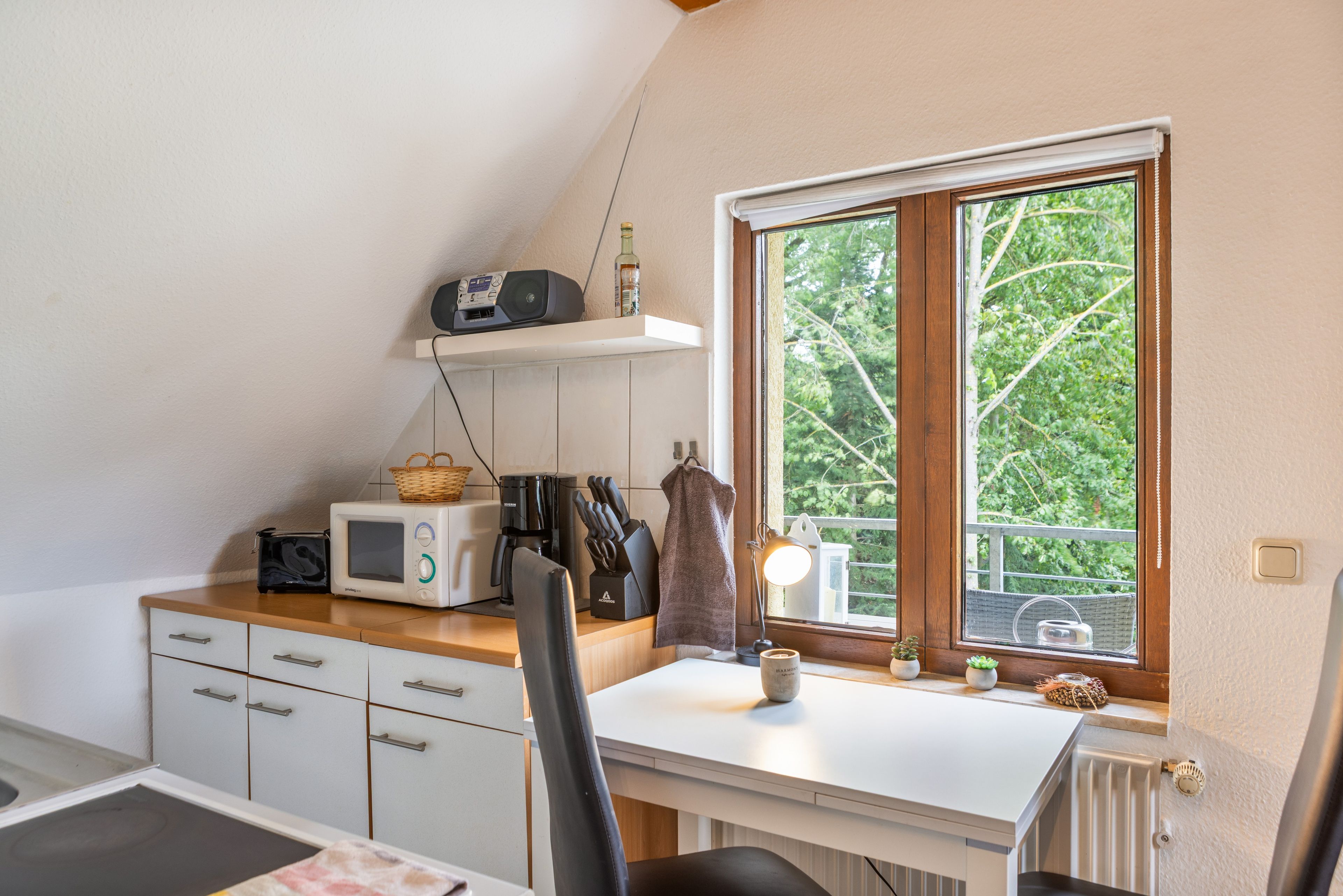 Una cocina con mostrador blanco, microondas, tostadora, cafetera y ventana con vista a árboles.