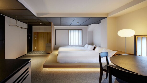 A hotel room with a bed, a desk, and a chair. There is a window with blinds, a lamp, and a door leading to another room.