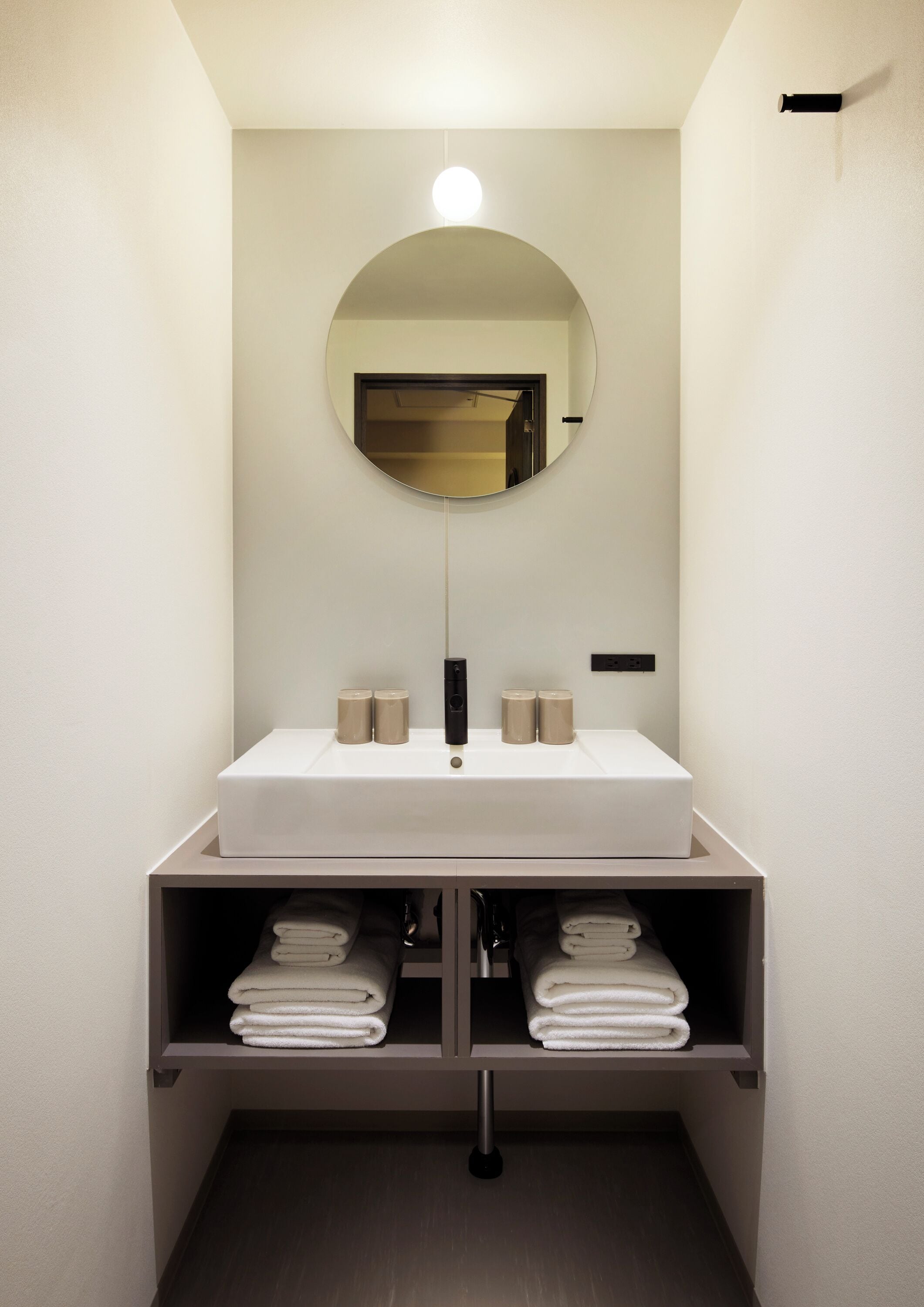 A modern bathroom with a white sink, a round mirror, and a shelf with folded towels.
