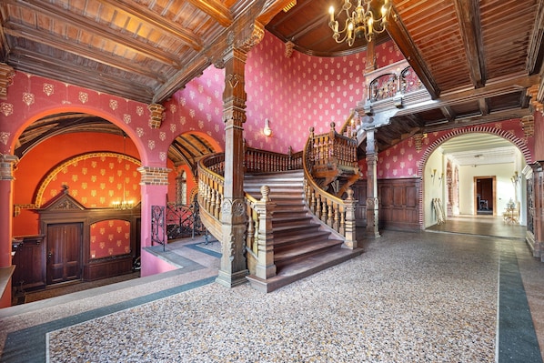 A grand staircase with ornate woodwork, a red patterned wall, and a chandelier.