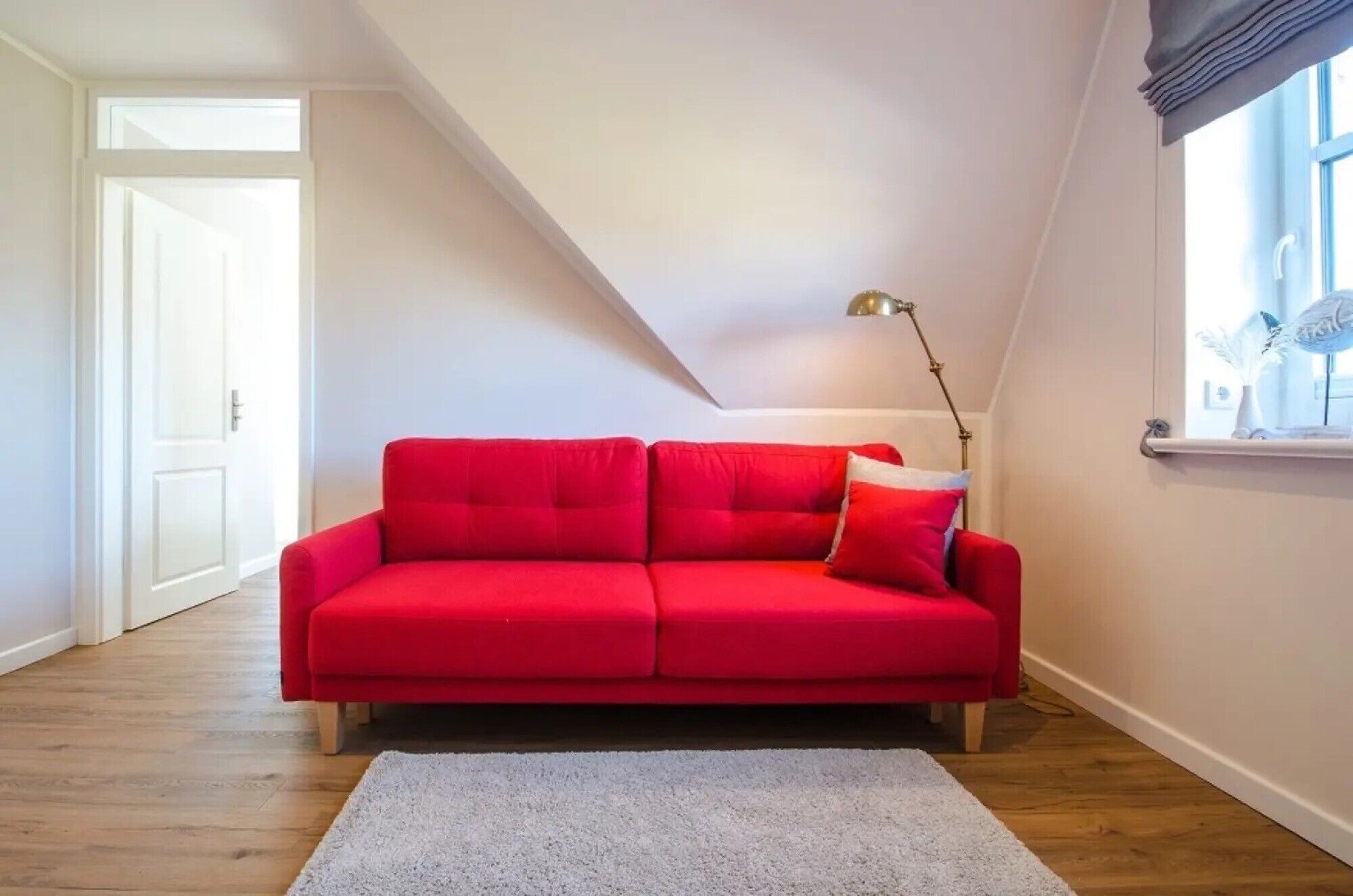 A red sofa in a room with wooden flooring, a white door, and a window with a curtain.
