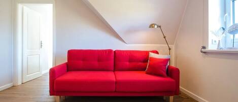 A red sofa in a room with wooden flooring, a white door, and a window with a curtain.