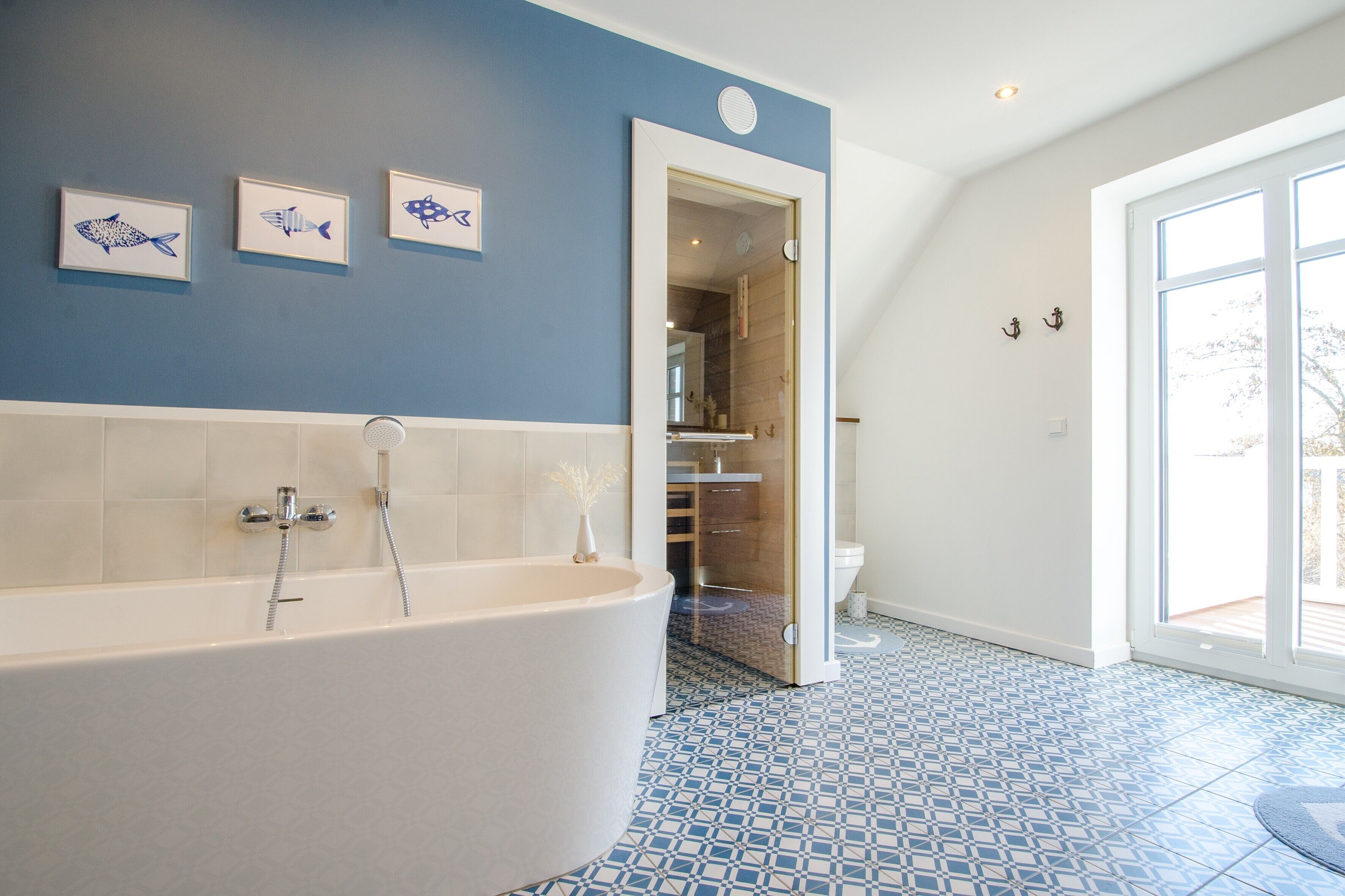 Een moderne badkamer met een vrijstaand bad, een douche en een toilet. Drie kunstwerken met vismotieven in lijsten en een groot raam zorgen voor natuurlijk licht.
