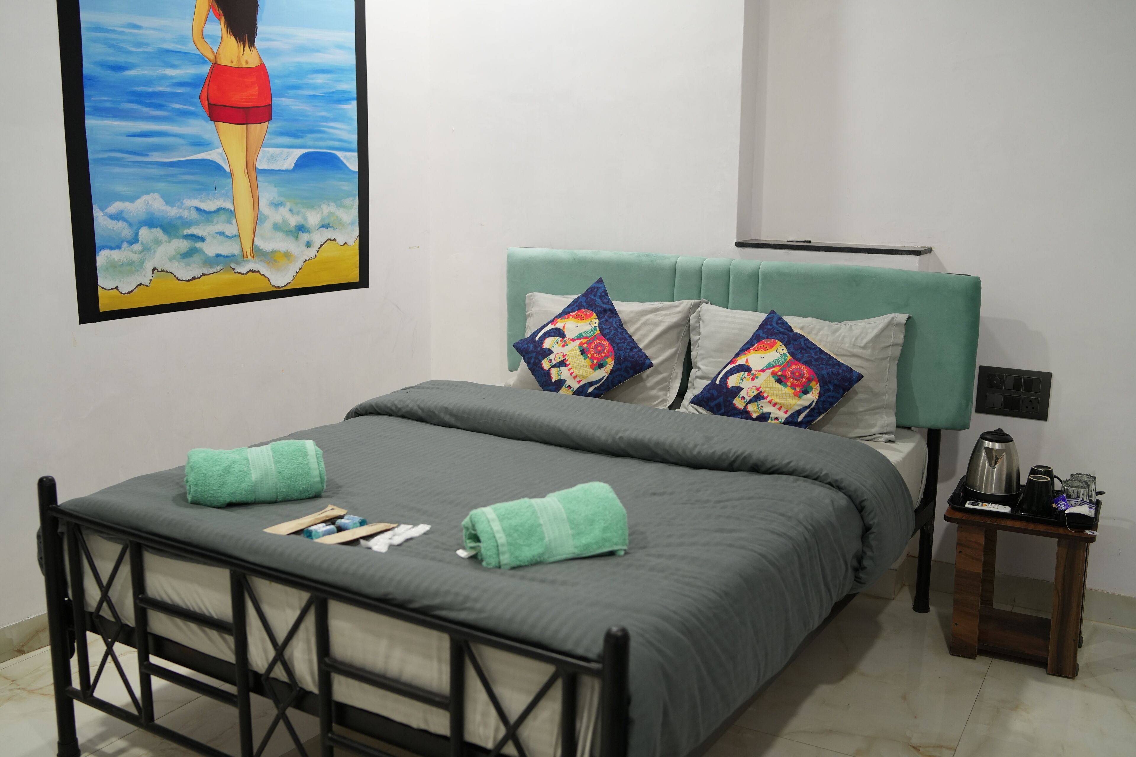 Ein ordentlich bezogenes Bett mit einer grauen Tagesdecke und zwei grünen Handtüchern, ein moosgrüner, gepolsterter Kopfteil und ein kleiner Nachttisch mit Wasserkocher und Fernbedienung.