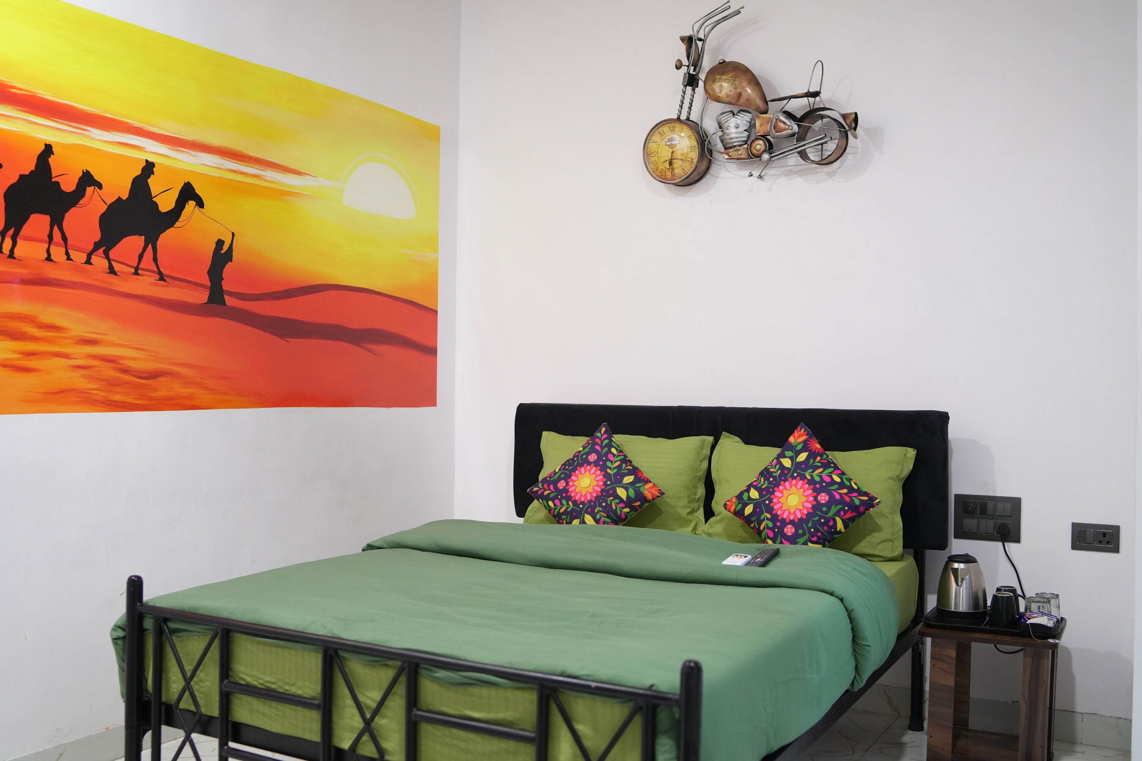 Un lit recouvert d’une couvre-lit verte et agrémenté de coussins colorés, une table de chevet avec une bouilloire et une télécommande, et un tableau mural représentant un paysage désertique avec des chameaux et une personne.