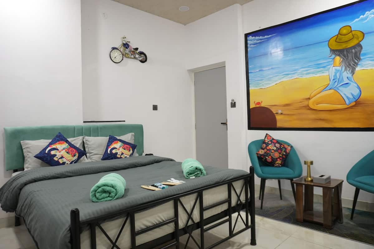 Une chambre à coucher avec un lit, des fauteuils turquoise, une petite table et un tableau représentant une femme sur une plage.