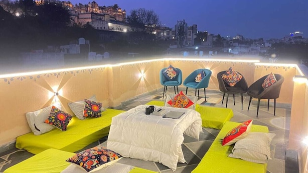 Una terrazza sul tetto con diversi letti e sedie, alcune con cuscini colorati, e una vista sulla città sullo sfondo.