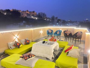 Une terrasse sur le toit, dotée de plusieurs lits et de chaises, dont certains sont agrémentés de coussins colorés, et offrant une vue sur le paysage urbain en arrière-plan.