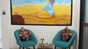 Una stanza con un grande quadro raffigurante una donna su una spiaggia, due poltrone color petrolio con cuscini decorati, un piccolo tavolino e un letto con una coperta verde.