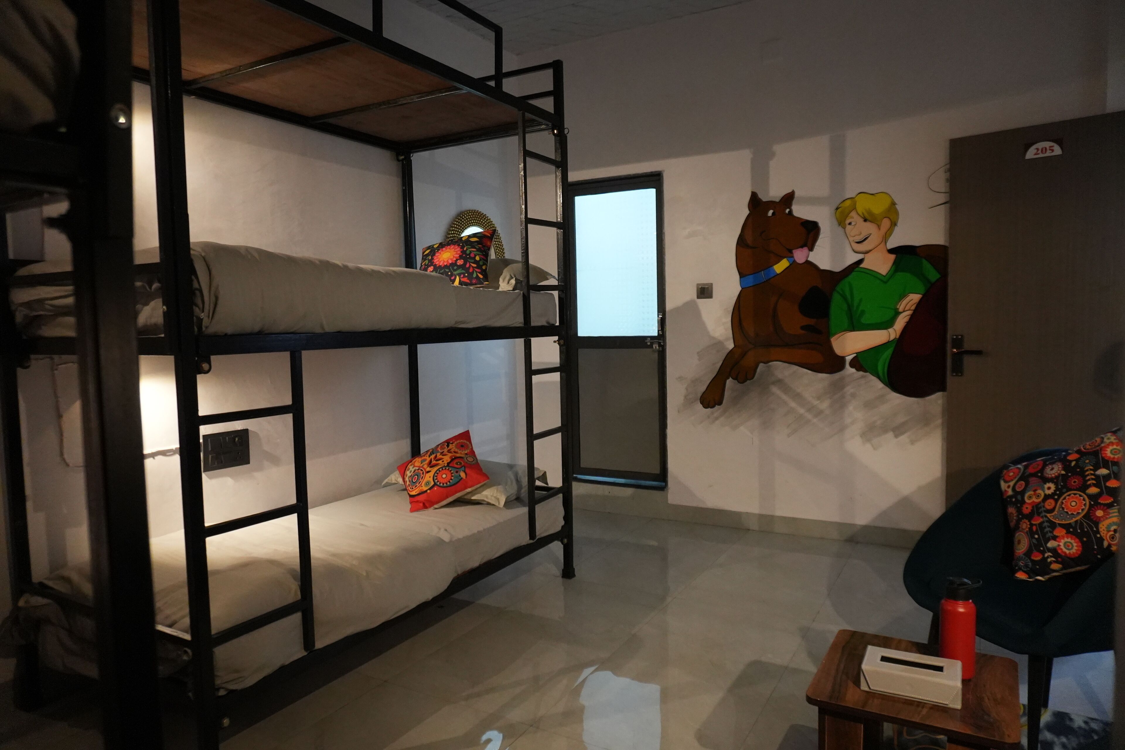 A bunk bed with a lower bunk, a small table with a red water bottle, and a wall with a cartoon character and a door marked with the number 205.
