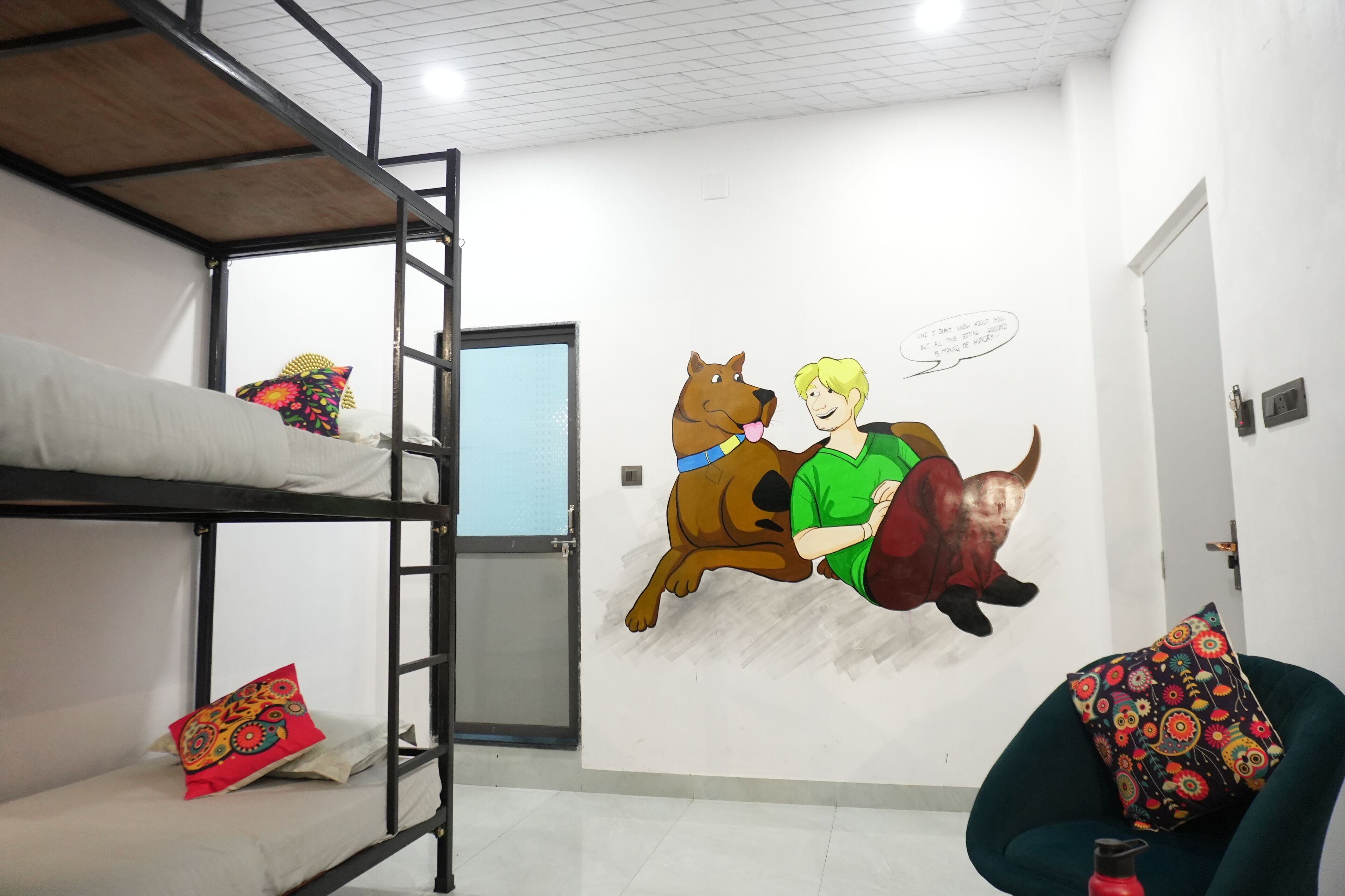 A bunk bed room with a mural of Scooby-Doo and a person, a door, and a chair with a colorful cushion.