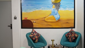 Una stanza con un quadro di grandi dimensioni a tema spiaggia, sedie color acquamarina con cuscini decorati e un tavolino con una lampada.
