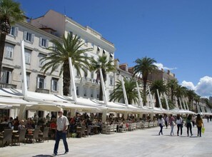 Un concurrido espacio al aire libre con cafetería, toldos blancos y palmeras, rodeado de edificios históricos.