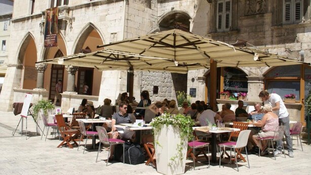 An outdoor café with tables and chairs, set against a historic stone building with arched windows and balconies.