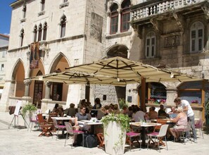 Een terras met tafels en stoelen, gelegen voor een historisch gebouw van steen met gebogen ramen en balkons.
