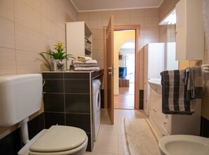 A compact bathroom with a toilet, bidet, and a sink. A towel rack is visible, and a door leads to a hallway.
