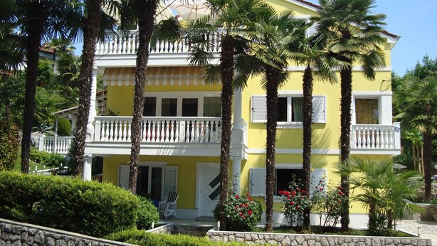 A two-story yellow building with a balcony, surrounded by palm trees and a stone wall.