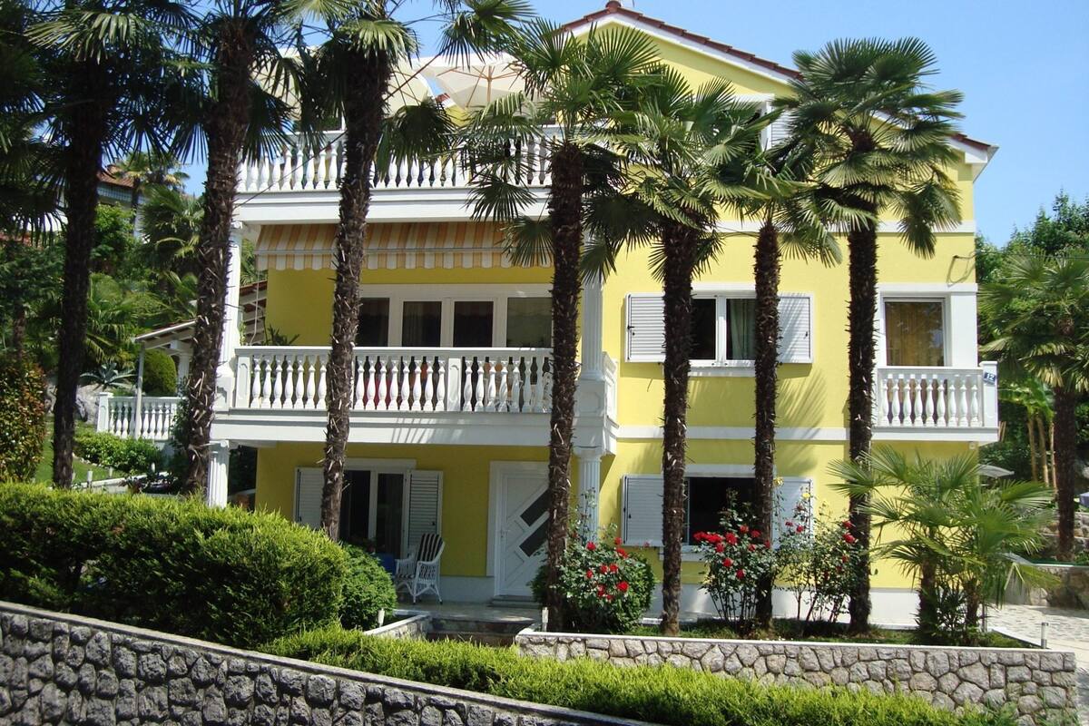 A two-story yellow building with a balcony, surrounded by palm trees and a stone wall.