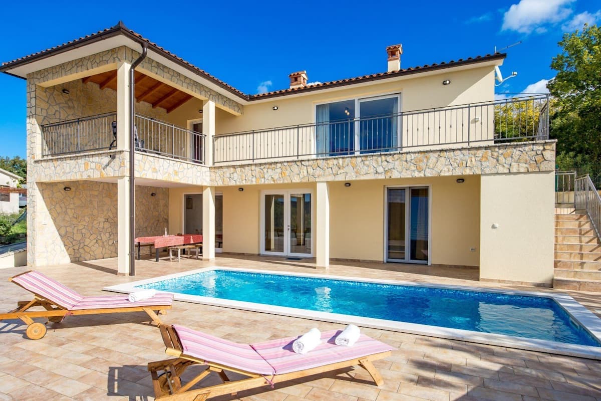 Una casa de dos pisos con piscina, mobiliario de exterior y fachada de piedra.