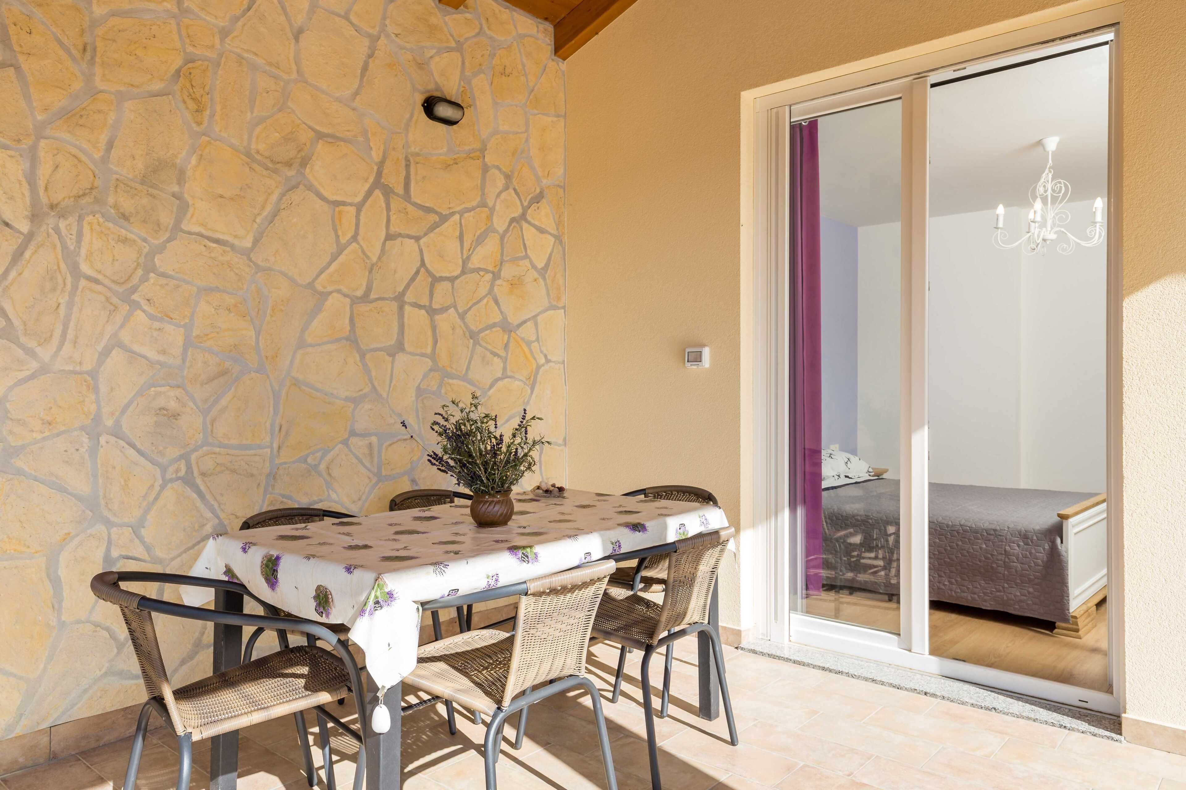 Un coin repas avec un mur en pierre, une table recouverte d’une nappe à motifs, des chaises en osier et une vue sur une chambre à coucher à travers une porte coulissante.