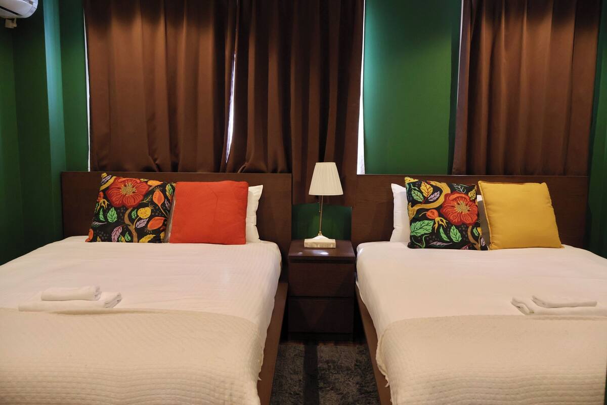 Two beds with colorful pillows, a nightstand with a lamp, and a green wall.