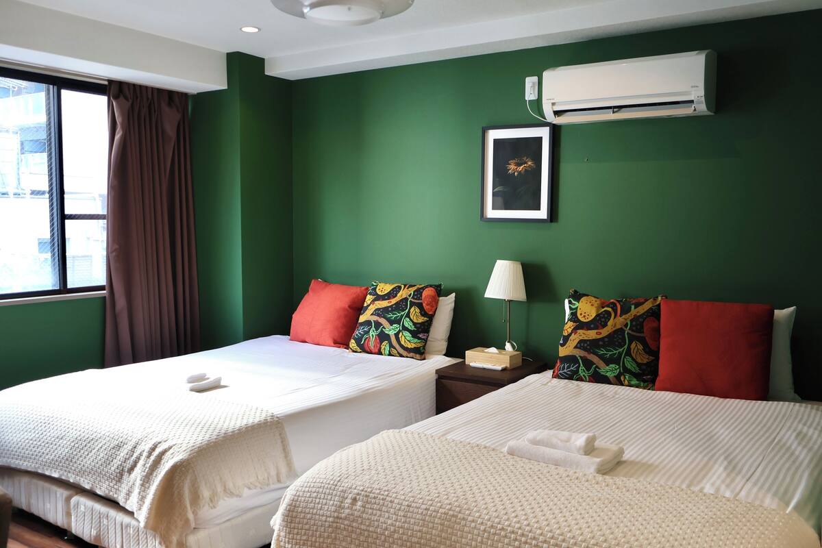 A hotel room with two beds, green walls, a window with curtains, an air conditioning unit, and a framed picture on the wall.