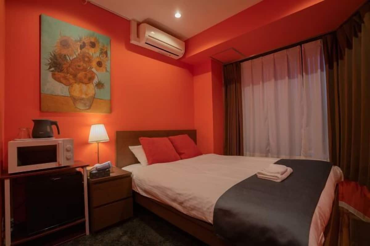A hotel room with a bed, bedside table, lamp, air conditioning unit, and a painting on the wall.