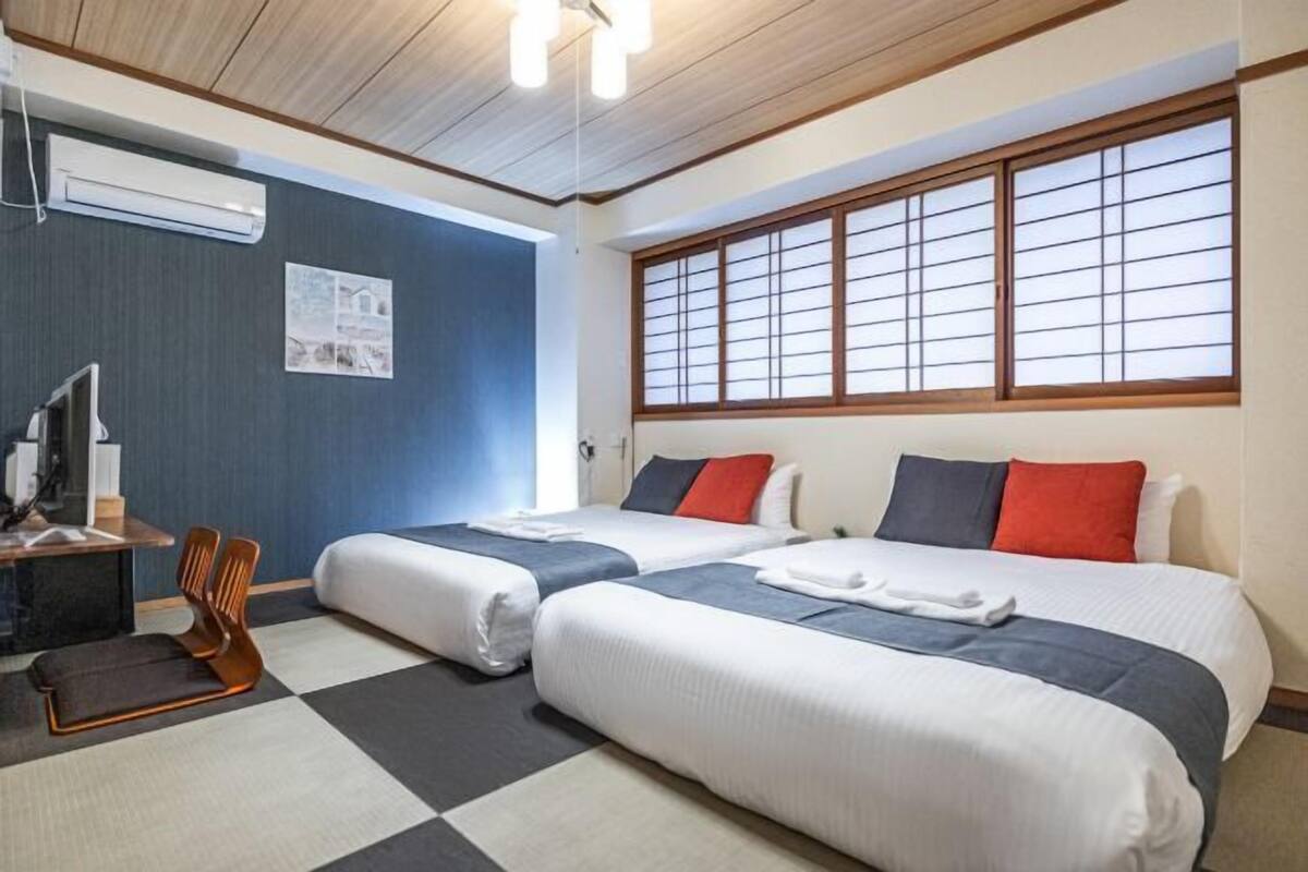 A hotel room with two beds, a desk, a chair, and a large window with traditional shutters.