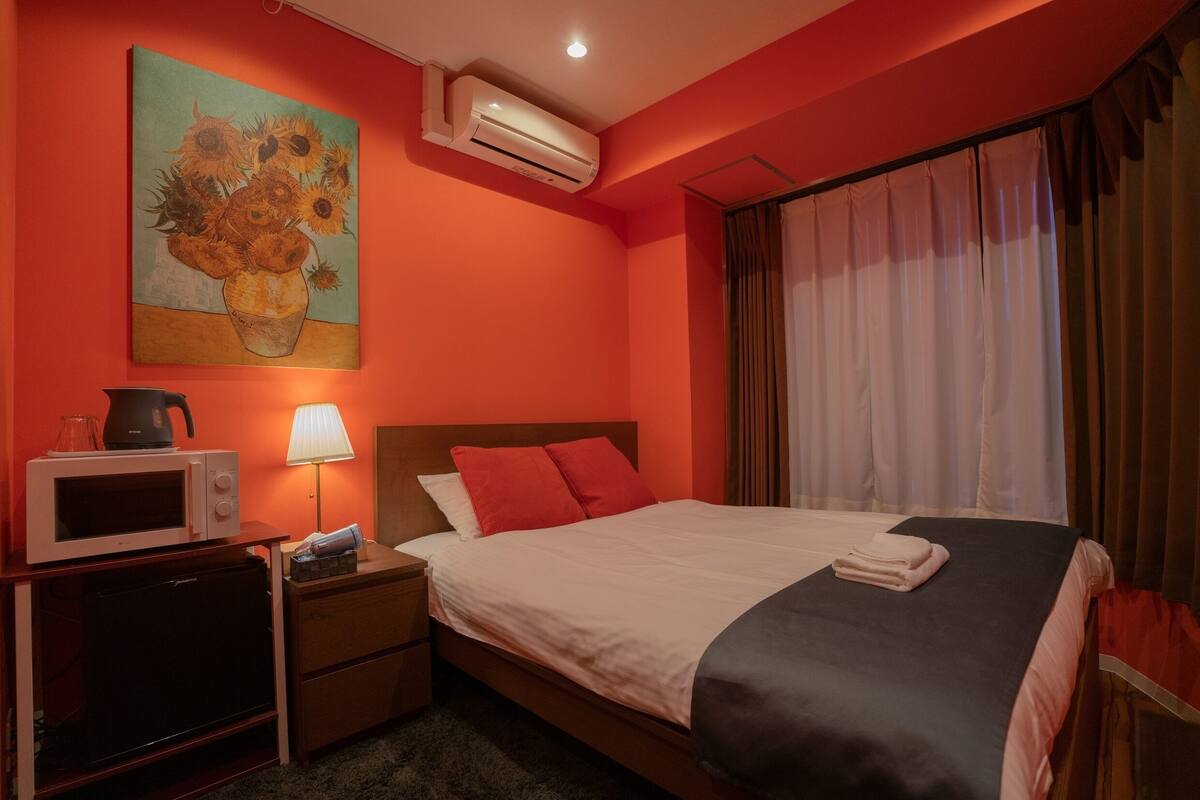 A hotel room with a bed, a nightstand, a microwave, an air conditioner, and a painting on the wall.
