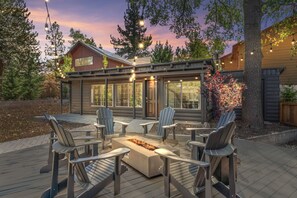 A cozy outdoor patio with Adirondack chairs and a fire pit, surrounded by wooden cabins with large windows and string lights. - Moonridge escape w/ hot tub & fire pit (Big Bear Lake)