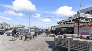 Una terraza de madera con mobiliario exterior, luces colgantes y vistas al puerto deportivo.