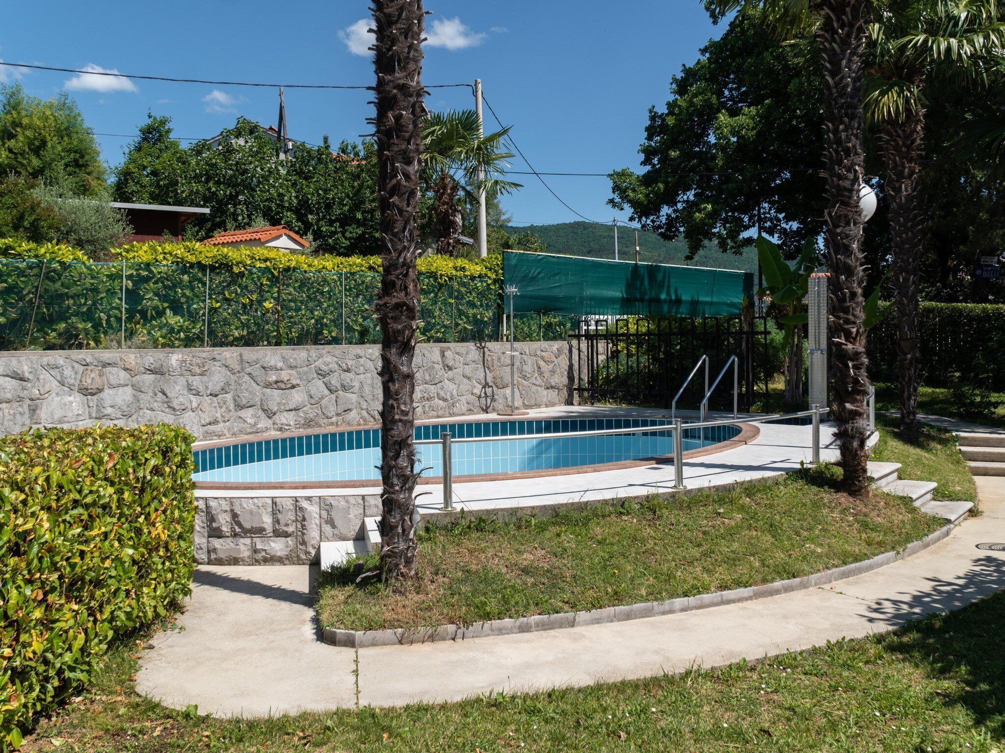 Una piscina con un muro de piedra y barandilla metálica, rodeada de césped y palmeras.