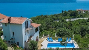 A two-story building with a swimming pool, surrounded by greenery and overlooking a body of water with mountains in the distance. - Charming Brsec Apartment With Shared Pool (Moscenicka Draga)