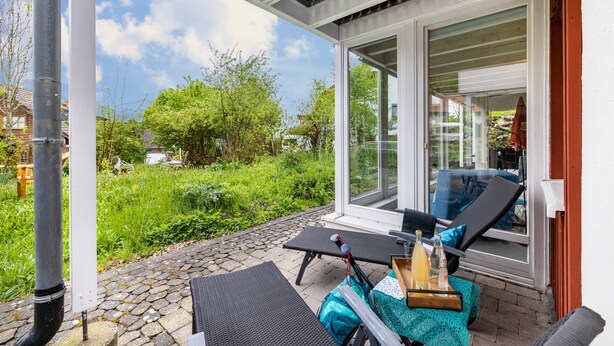 A covered outdoor seating area with lounge chairs, a table with a drink, and a view of greenery and a building. - Apartment 'Untere Ferienwohnung' with Private Terrace, Shared Garden and Wi-Fi (Meßstetten)