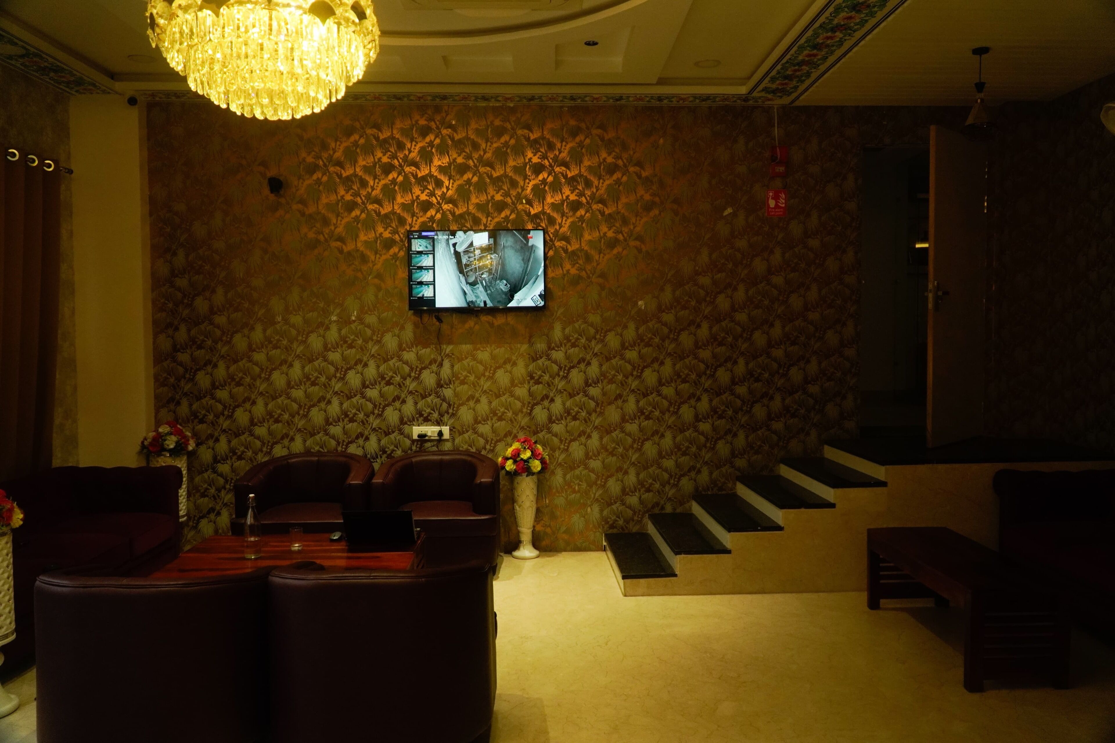 A room with a patterned wallpaper, a chandelier, a flat-screen TV mounted on the wall, a sofa, a coffee table, and a staircase leading to an upper level.