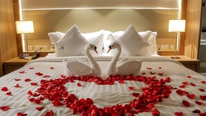 Quarto de hotel com uma cama, almofadas em formato de cisne e um arranjo de pétalas de rosa em formato de coração sobre a cama.