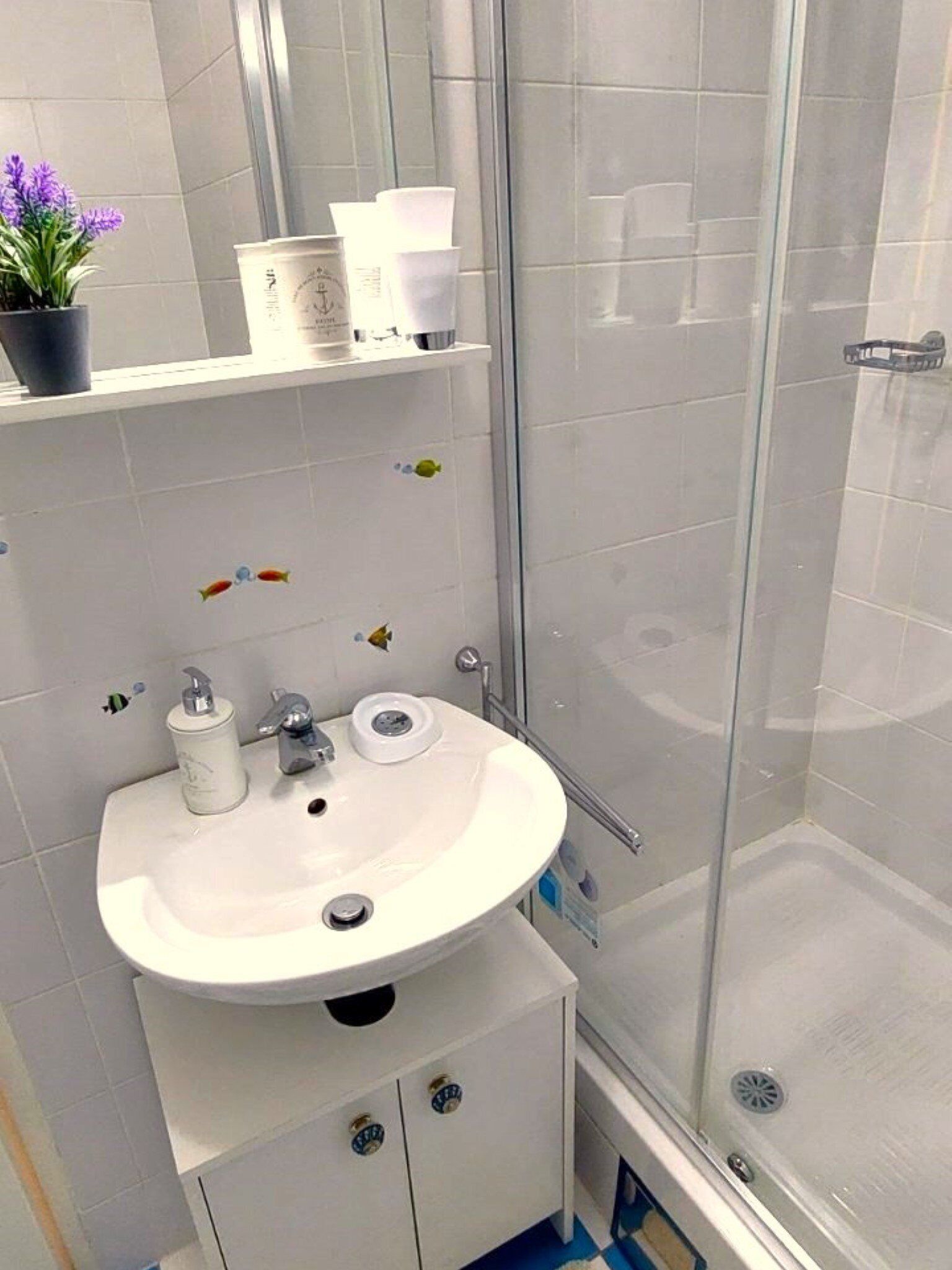 Une salle de bain avec un lavabo blanc, un miroir, un espace douche et une étagère contenant des articles de toilette.