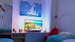 Een slaapkamer met een bed, een bureau met een computer, een televisie en een schilderij van zeilboten.