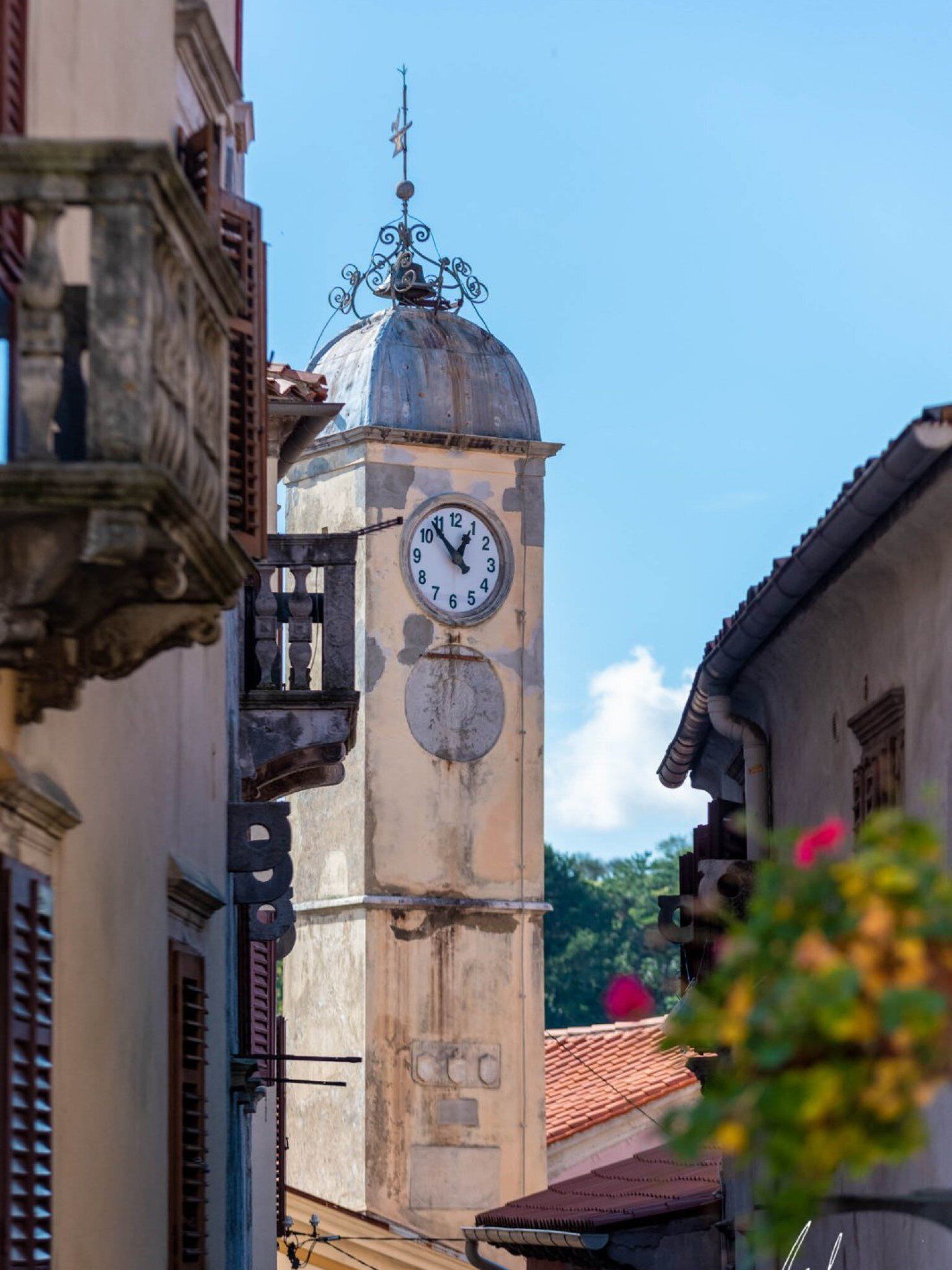 A historic clock tower with a weather vane on top, surrounded by traditional buildings.