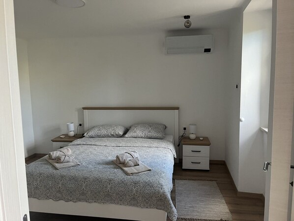 A bedroom with a bed, two bedside tables, a nightstand, a wall-mounted air conditioner, and a door.