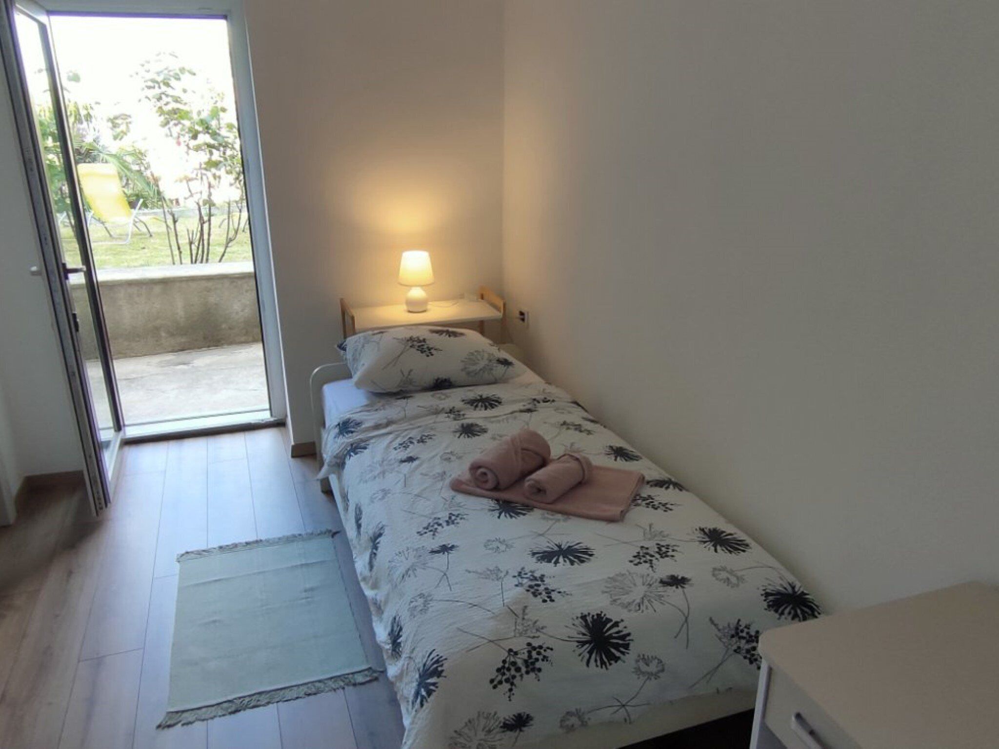 Une petite chambre avec un lit simple, une couvre-lit à motifs floraux, une lampe de chevet et une vue sur l’extérieur grâce à une porte coulissante.