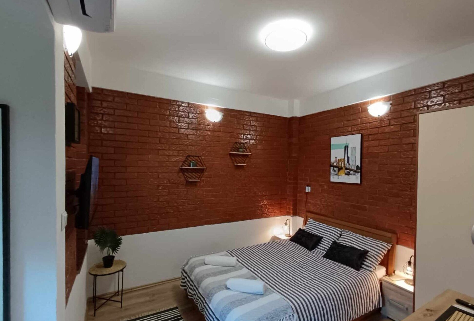 A bedroom with a brick wall, a bed with striped bedding, a small bedside table with a lamp, and a picture on the wall.