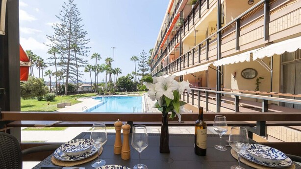 A dining area with a table set for two, overlooking a pool and a landscaped garden. - Sea Breeze Frontline Apartment (Los Cristianos)