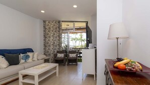 A modern living room with a sofa, coffee table, and a dining area with chairs. A fruit bowl is on a wooden table. - Sea Breeze Frontline Apartment (Los Cristianos)