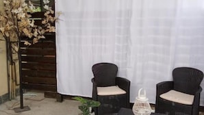 Two wicker chairs with cushions, a white lantern, and a potted plant in a patio area with a white curtain backdrop. - LaBandaBassottiFerrara  (Monestirolo)