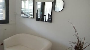 A white sofa in a room with a window, a wall-mounted mirror, and a vase with dried plants. - LaBandaBassottiFerrara  (Monestirolo)