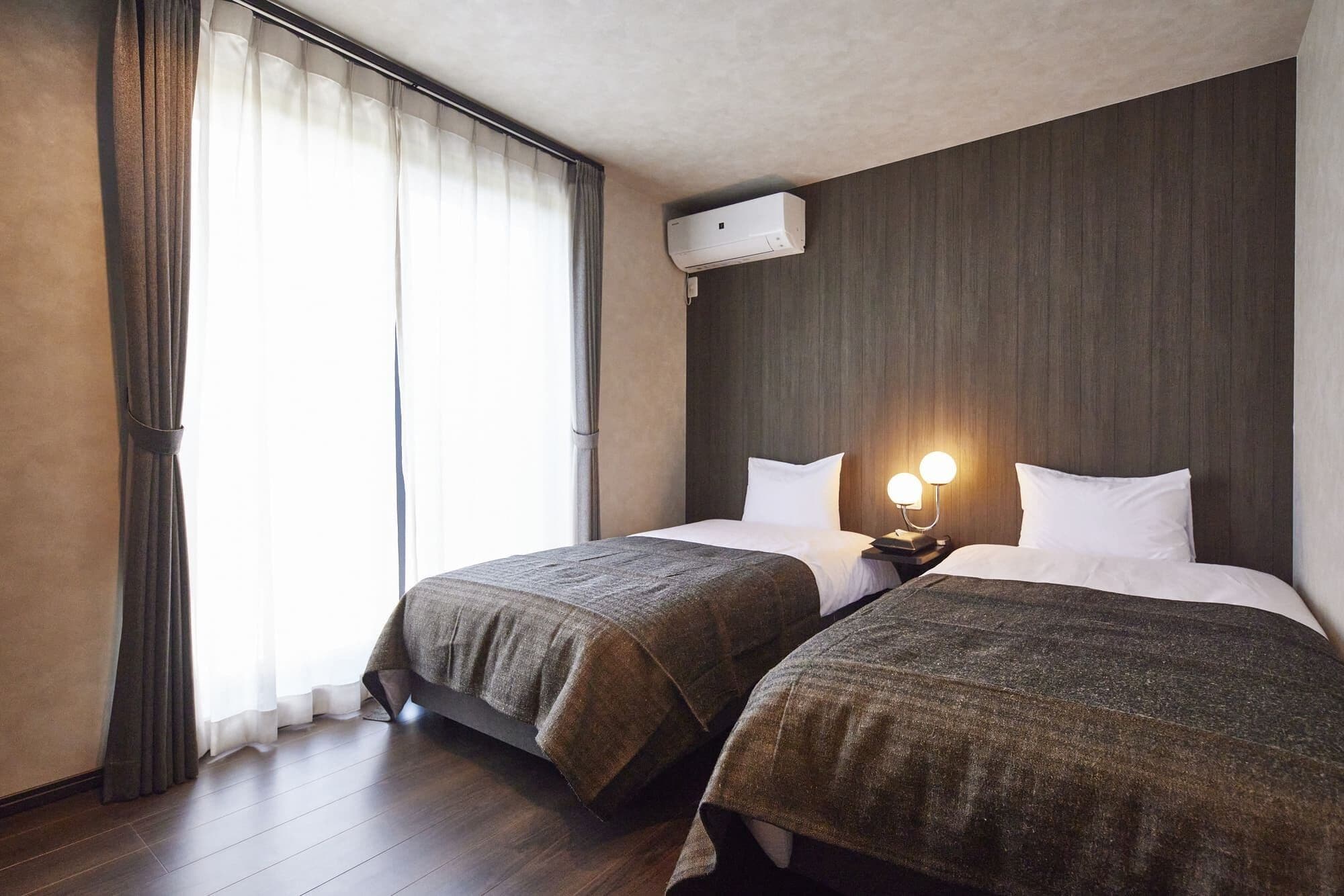 A hotel room with two beds, a window with curtains, an air conditioning unit, and a bedside lamp.