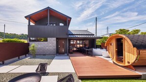 Una casa moderna de dos pisos con una sauna de madera, un patio con mobiliario exterior y un cielo despejado.