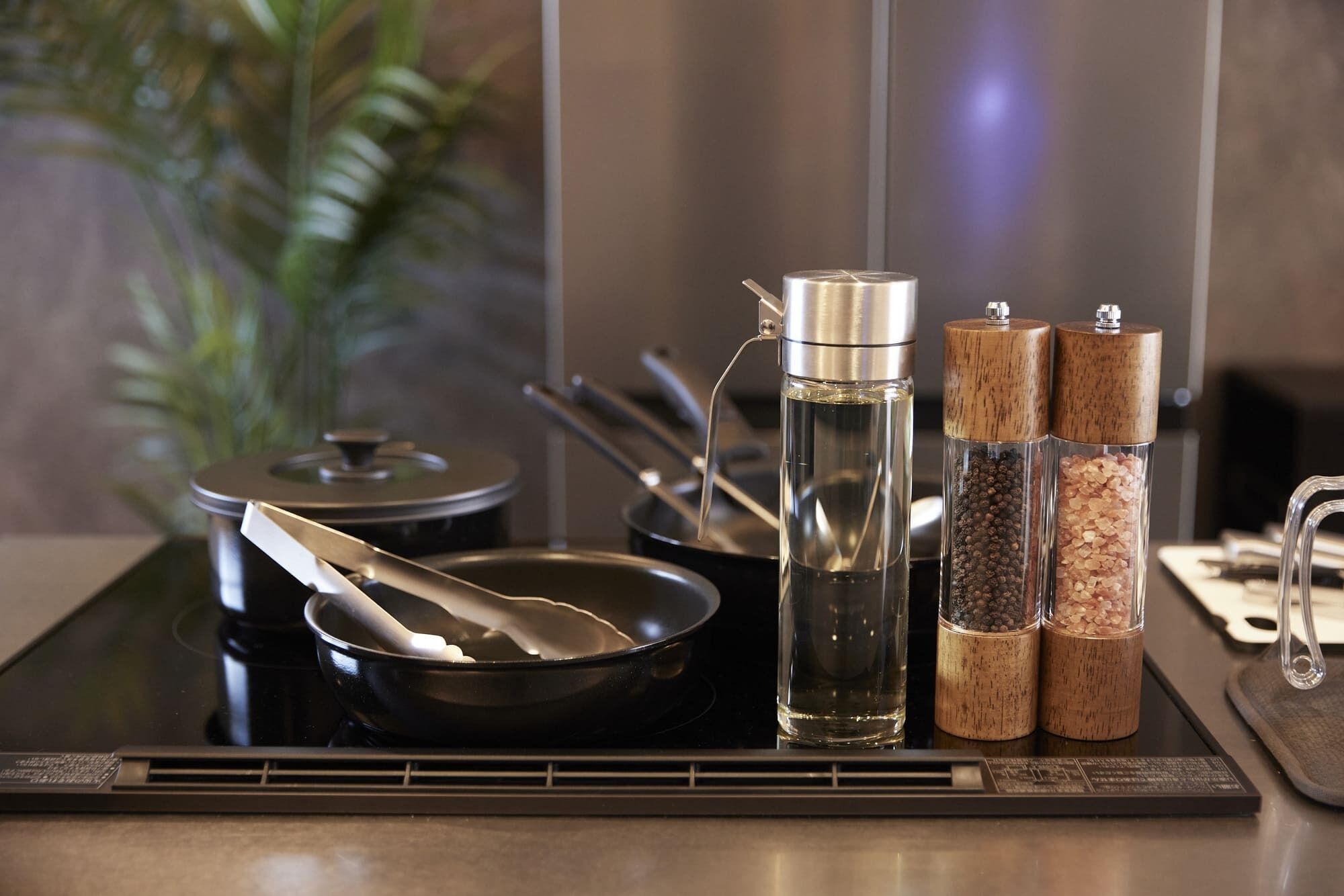 Un plan de travail de cuisine avec une plaque de cuisson à induction noire, une bouilloire en acier inoxydable, une poêle et deux pots à épices dotés de couvercles en liège.